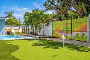 A backyard with a pool, artificial grass, a fence with a palm tree mural, and a flagpole. - Backyard Paradise • Grill • Game Room • Putt Putt (Palm Springs)