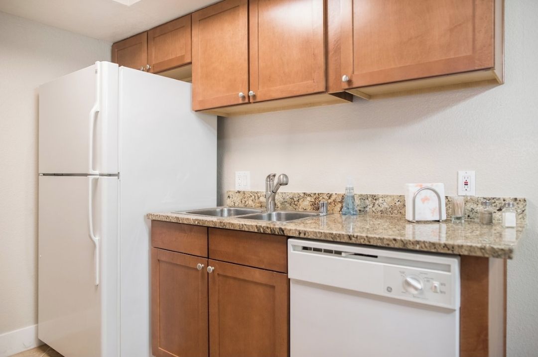 A kitchen with wooden cabinets, a white refrigerator, a granite countertop, a stainless steel sink, and a dishwasher.