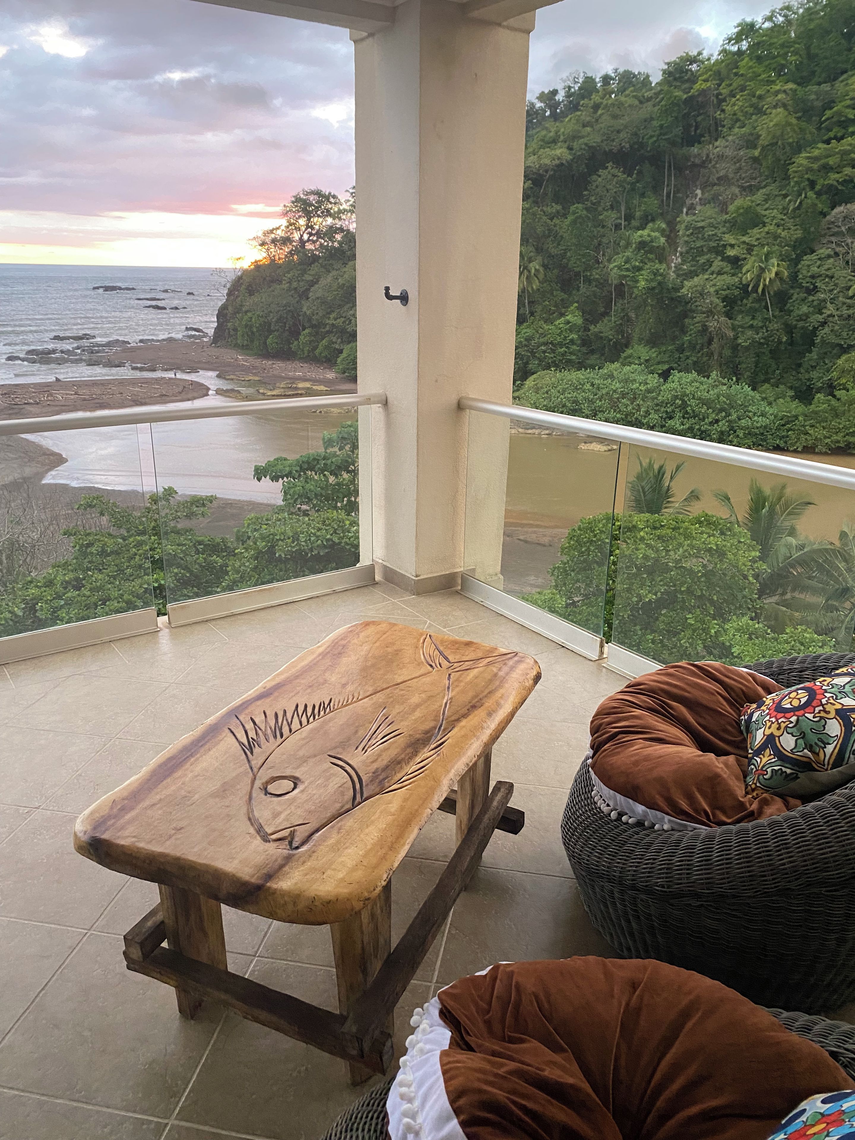 A balcony with a wooden bench and a wicker chair, overlooking a beach and forested area.