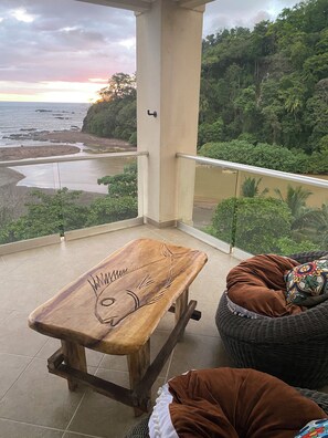 Un balcon avec un banc en bois et une chaise en osier, donnant sur une plage et une zone boisée.