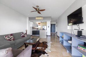 A modern living room with a grey sofa, a coffee table, and a cowhide rug. There is a kitchen in the background with a refrigerator and a ceiling fan. - Luxury Oceanfront 2-bedroom condo in enchanting Jacó on the Beach!!! (Jacó)
