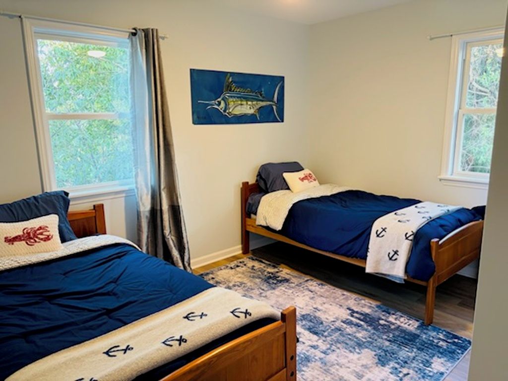 A bedroom with two single beds, a blue and white patterned rug, a painting of a fish on the wall, and windows with curtains.