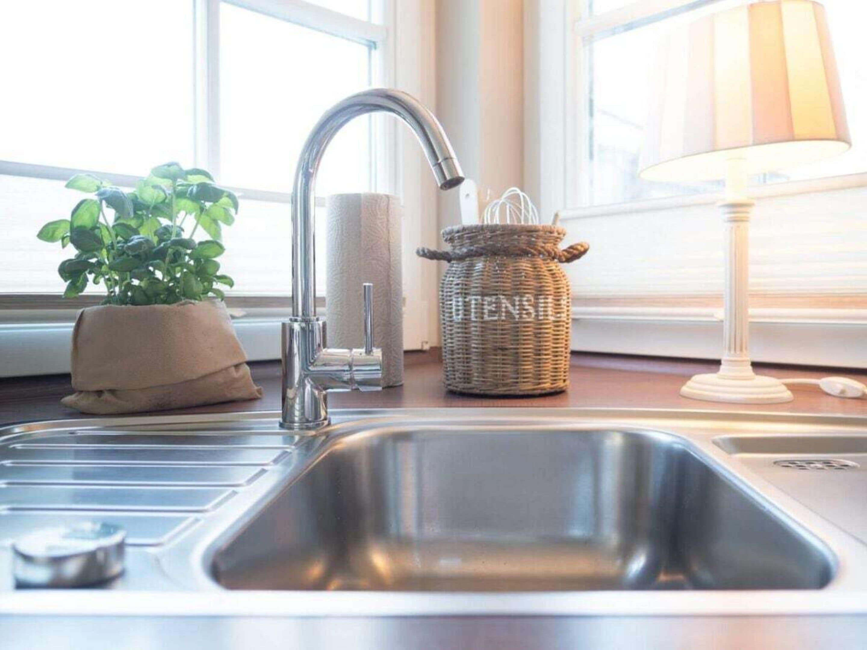 A kitchen sink with a modern faucet, a potted plant, a wicker basket labeled 'UTENSIL,' and a lamp on the countertop.