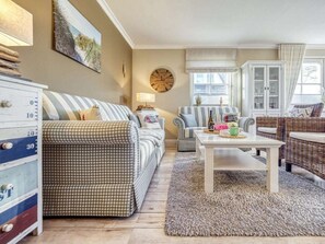 A cozy living room with a checkered sofa, a coffee table with decorative items, a clock on the wall, and a window with curtains. - House Kranich-kate (Zingst)