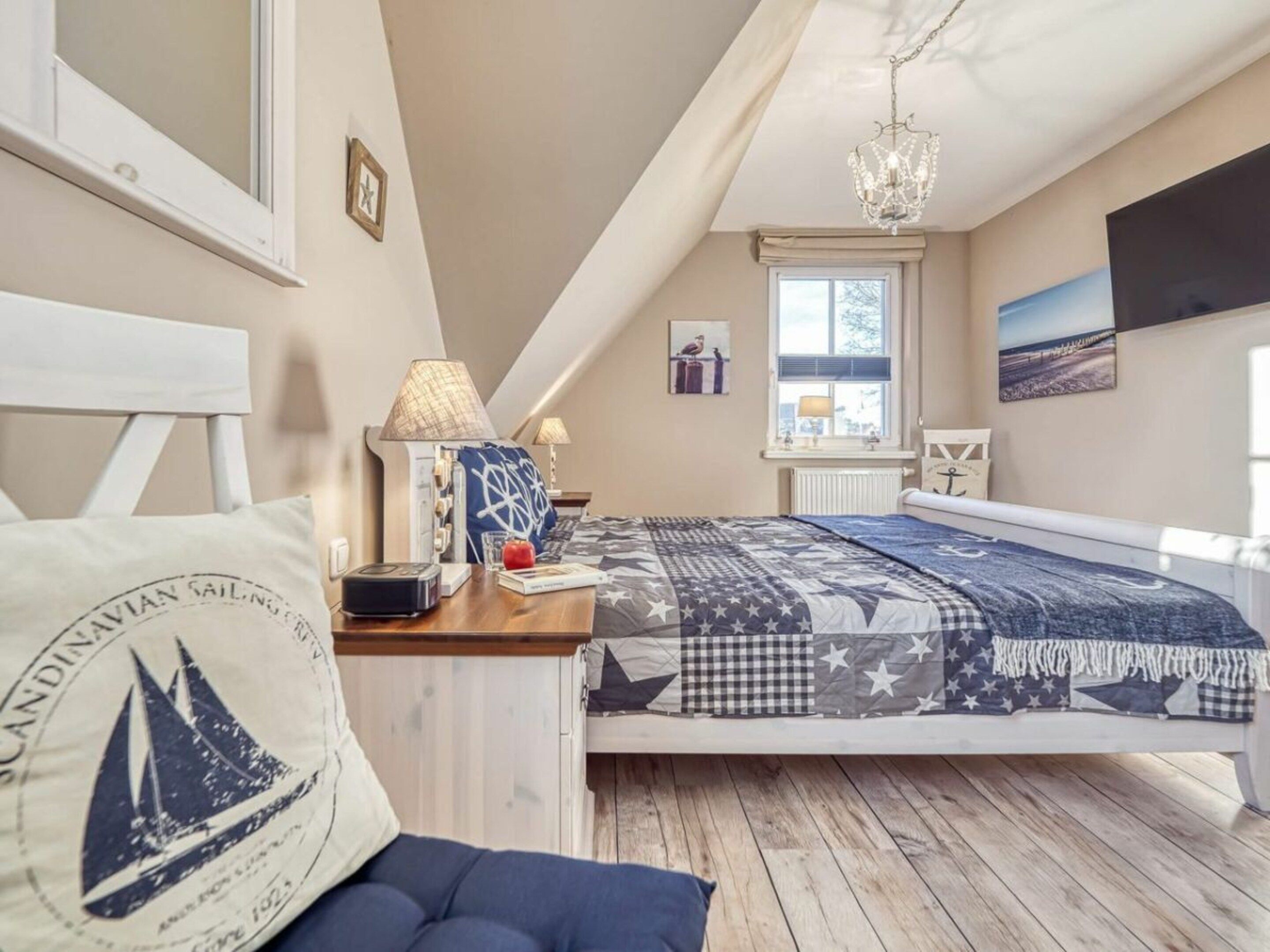 Un dormitorio con techo inclinado, una cama con colcha estampada, una mesita de noche con lámpara, una ventana con persianas y un televisor fijado a la pared.