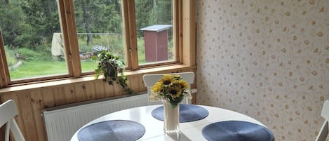 A dining area with a round table, four chairs, and a window with a view of greenery and a shed.