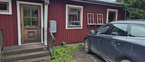 A red wooden house with a front door, a small porch, and a car parked in front.