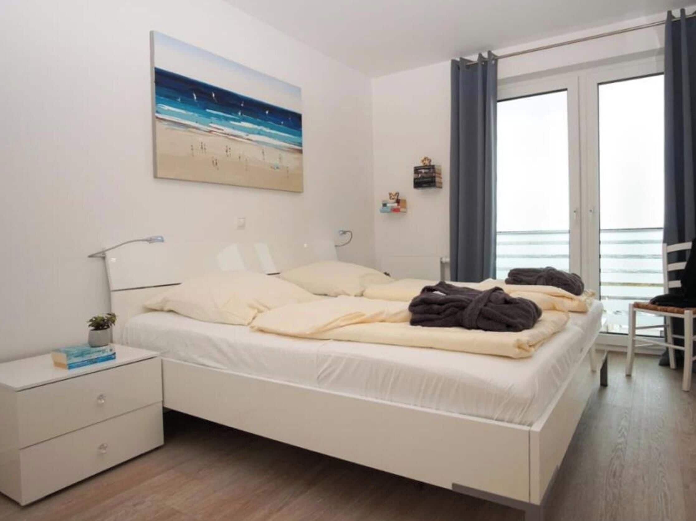 Un dormitorio con cama, mesitas de noche, un cuadro, una ventana con cortinas y vista al mar.
