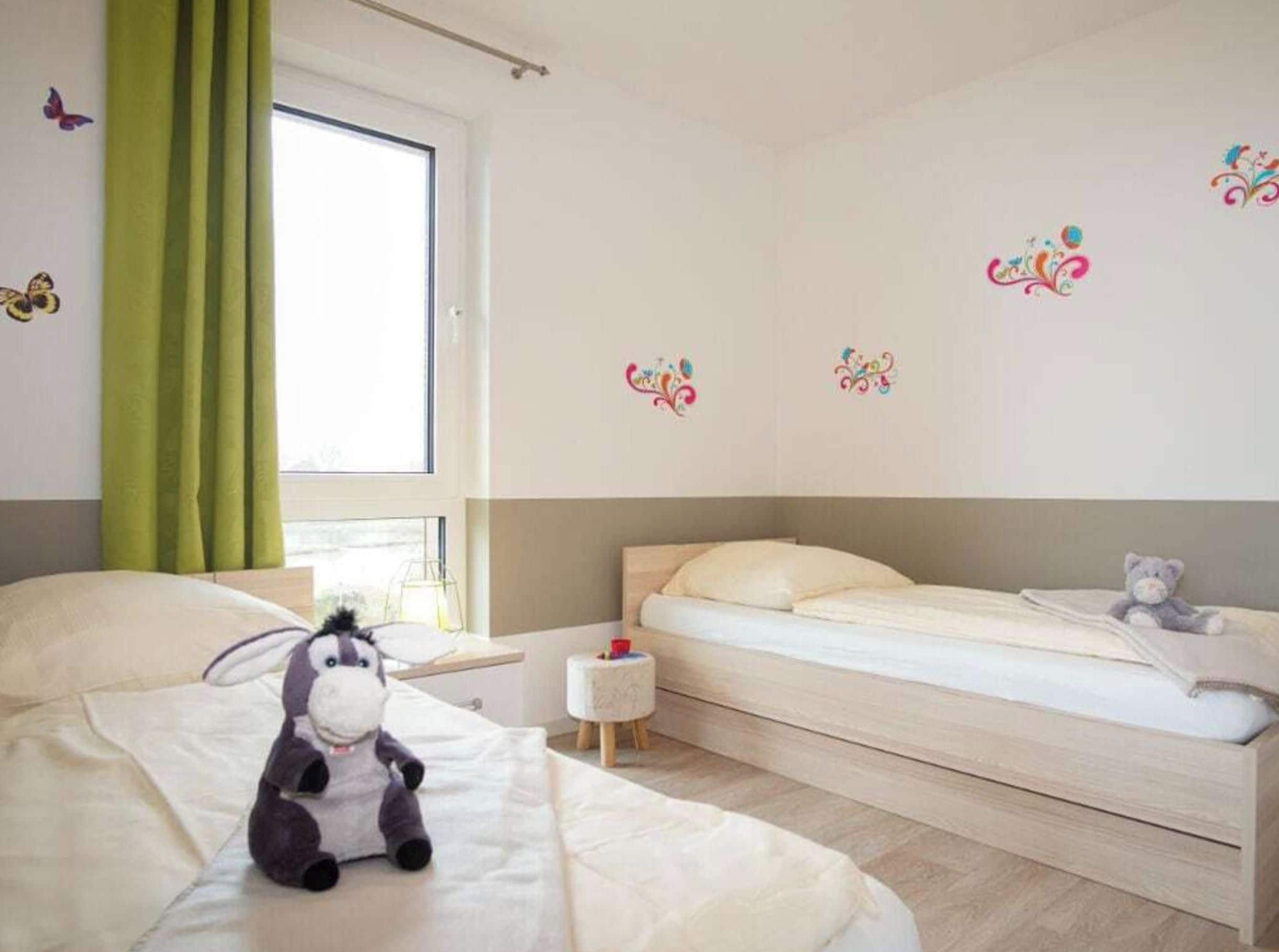 Een slaapkamer met twee bedden, een raam met gordijnen, decoratieve muurstickers en een pluchen speelgoed op een van de bedden.