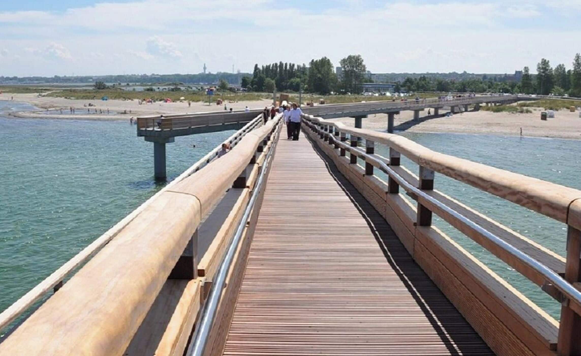 Ein hölzerner Steg führt zu einem Strand mit freiem Blick auf das Wasser und entfernte Gebäude.