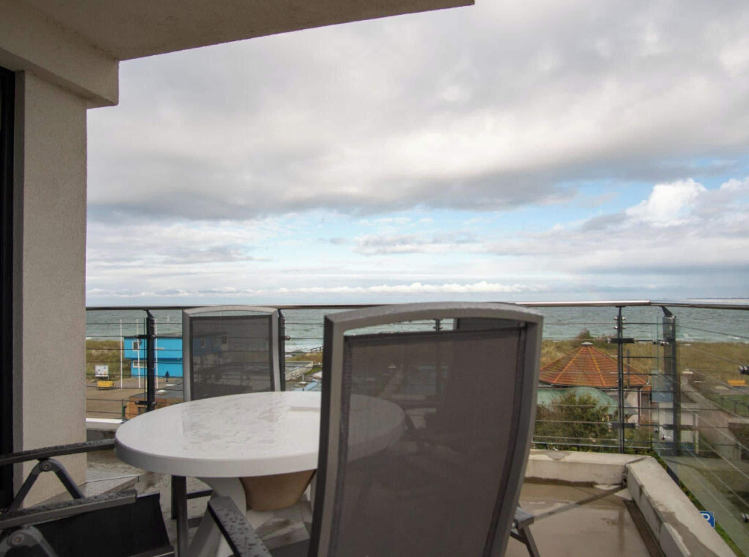 Ein Balkon mit rundem Tisch und Stühlen, mit Blick auf eine Küstenlandschaft mit Gebäuden und Meer.
