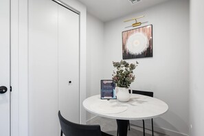 Ein modernes Zimmer mit einer weißen Tür, einem runden Tisch mit Blumenvase, einem begrüßungsschild im Bilderrahmen und einem an der Wand hängenden abstrakten Gemälde.