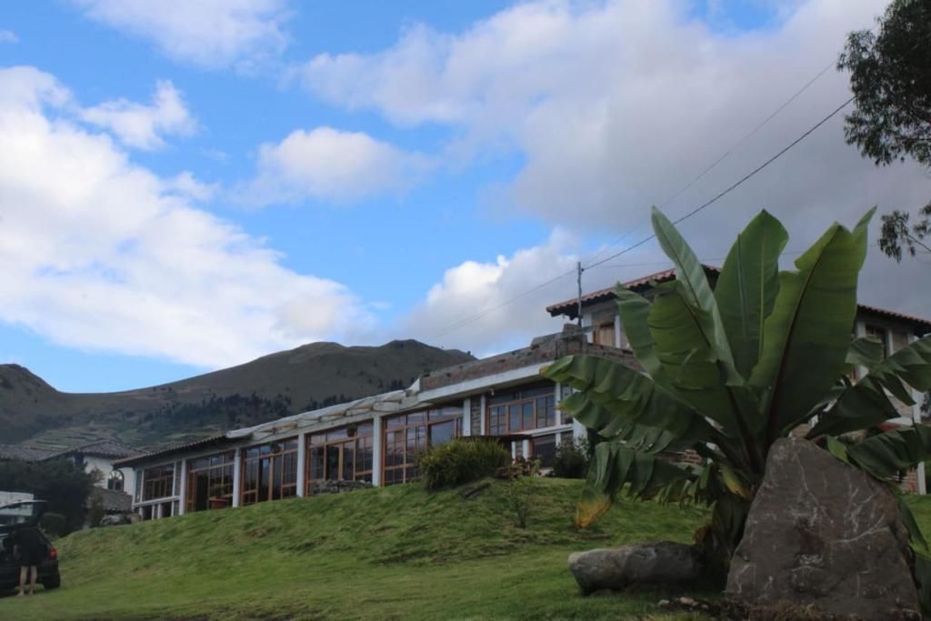 Un edificio con amplias ventanas y techo inclinado, rodeado de vegetación y colinas al fondo.