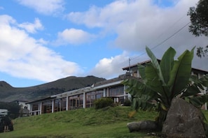 Un edificio con amplias ventanas y techo inclinado, rodeado de vegetación y colinas al fondo.