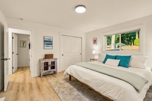Un dormitorio con cama, ventana con vistas, una puerta y un mueble pequeño.