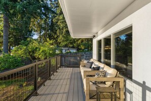 A wooden deck with a sofa and a table, surrounded by a metal railing and a view of trees and a building. - The Heron at Black Pearl | Dyes Inlet Retreat (Bremerton)