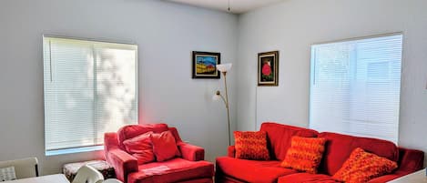 Una sala de estar con un sofá rojo, un ventilador de techo y dos ventanas con persianas.