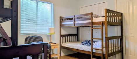 A bunk bed room with a desk and chair, a window with blinds, and a door.