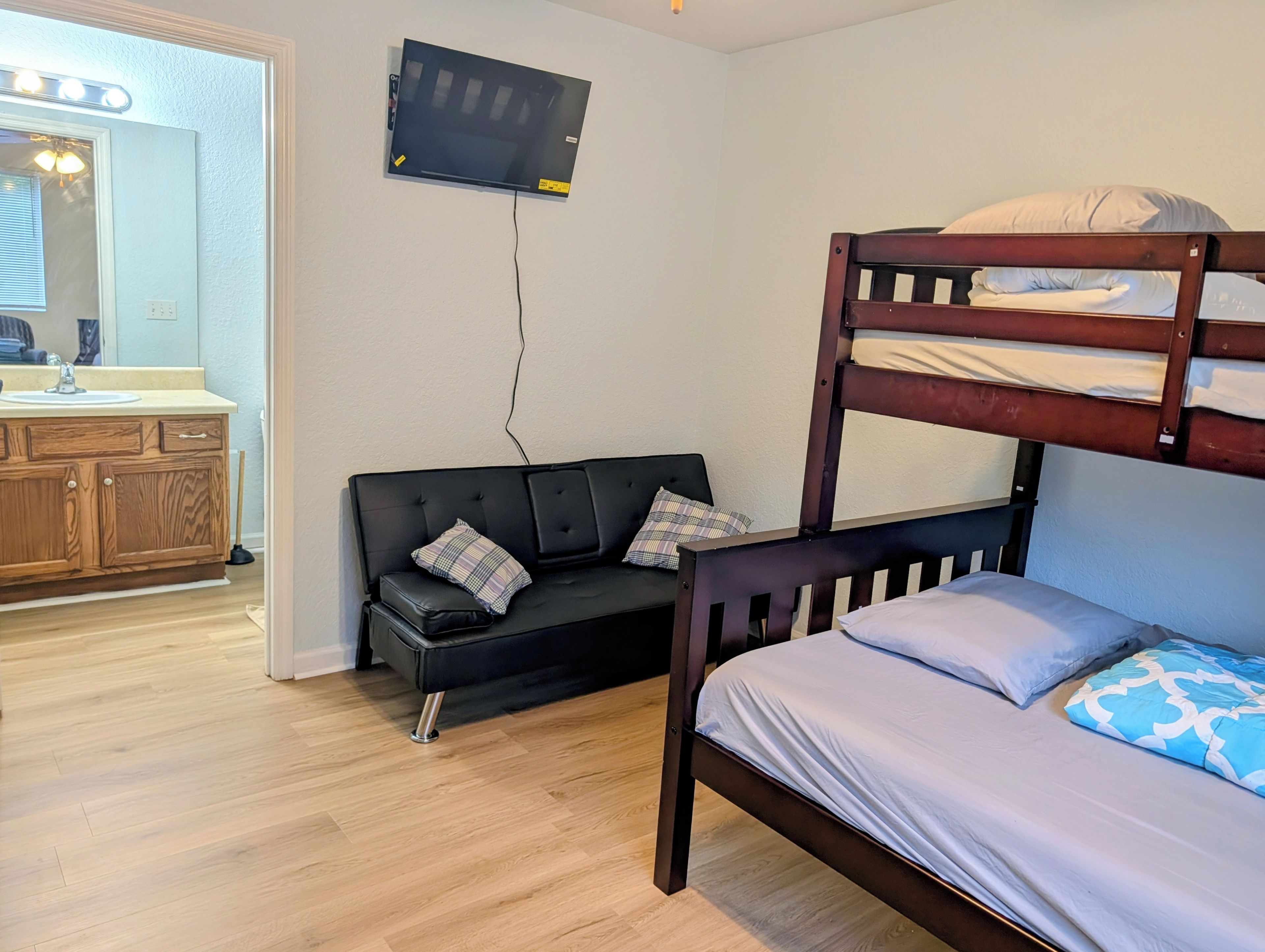 A room with a bunk bed, a sofa with pillows, a wooden cabinet, a television mounted on the wall, and a bathroom with a mirror and sink.