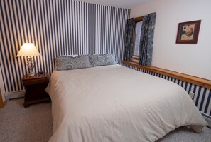 A neatly made bed with a white comforter and patterned pillows, a bedside lamp, and a striped wall with a framed picture. - 1BR Ski-in/Ski-out | Free Spring House Pool Access (Ludlow)