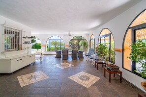 Un espace intérieur spacieux doté d’un sol carrelé, de coins salon et de grandes fenêtres offrant une vue sur la verdure.