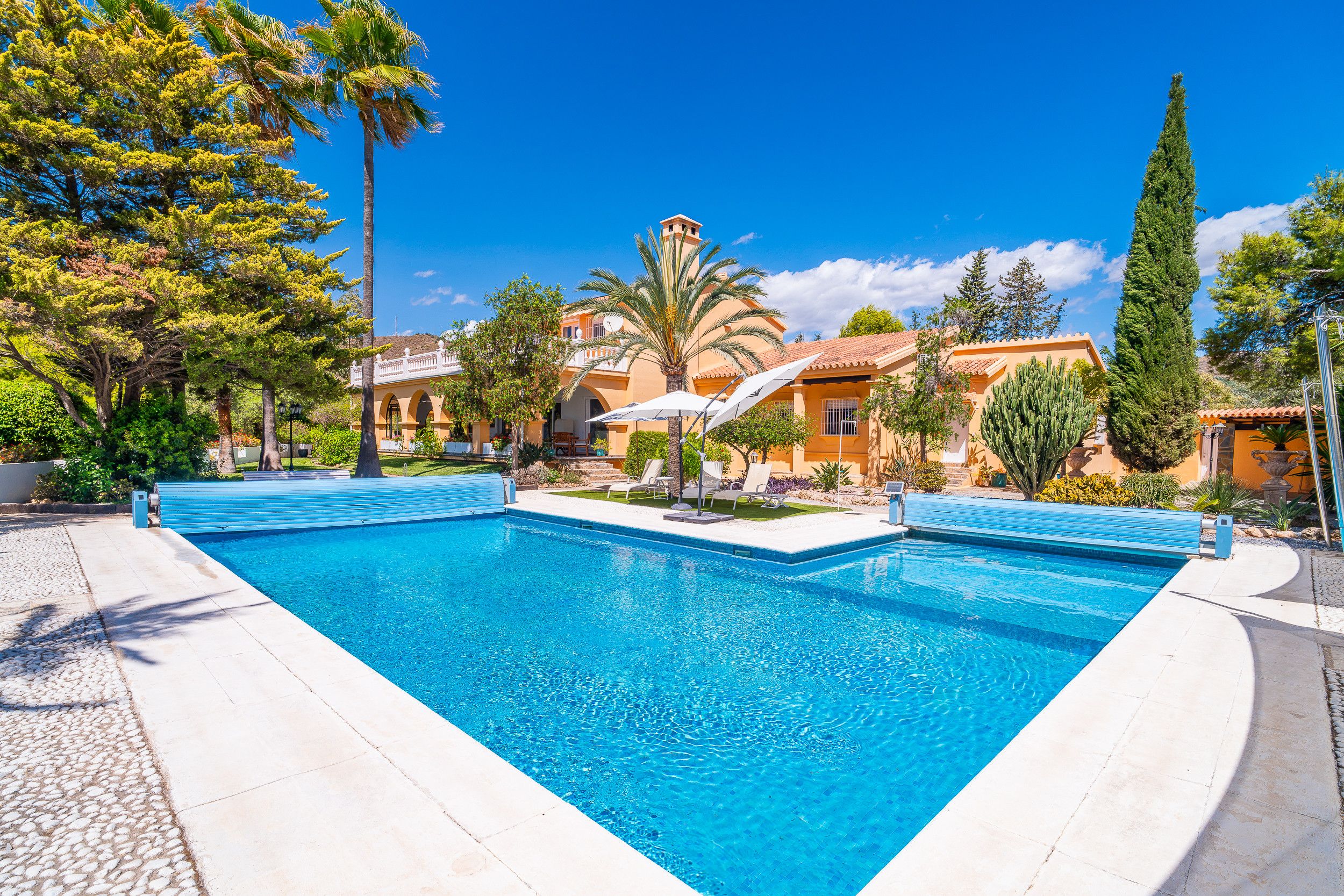 Une piscine à l’eau d’un bleu cristallin, entourée d’une terrasse dallée. On y trouve des chaises longues et des parasols, et la piscine est située juste à côté d’un bâtiment dont le toit est recouvert de tuiles.