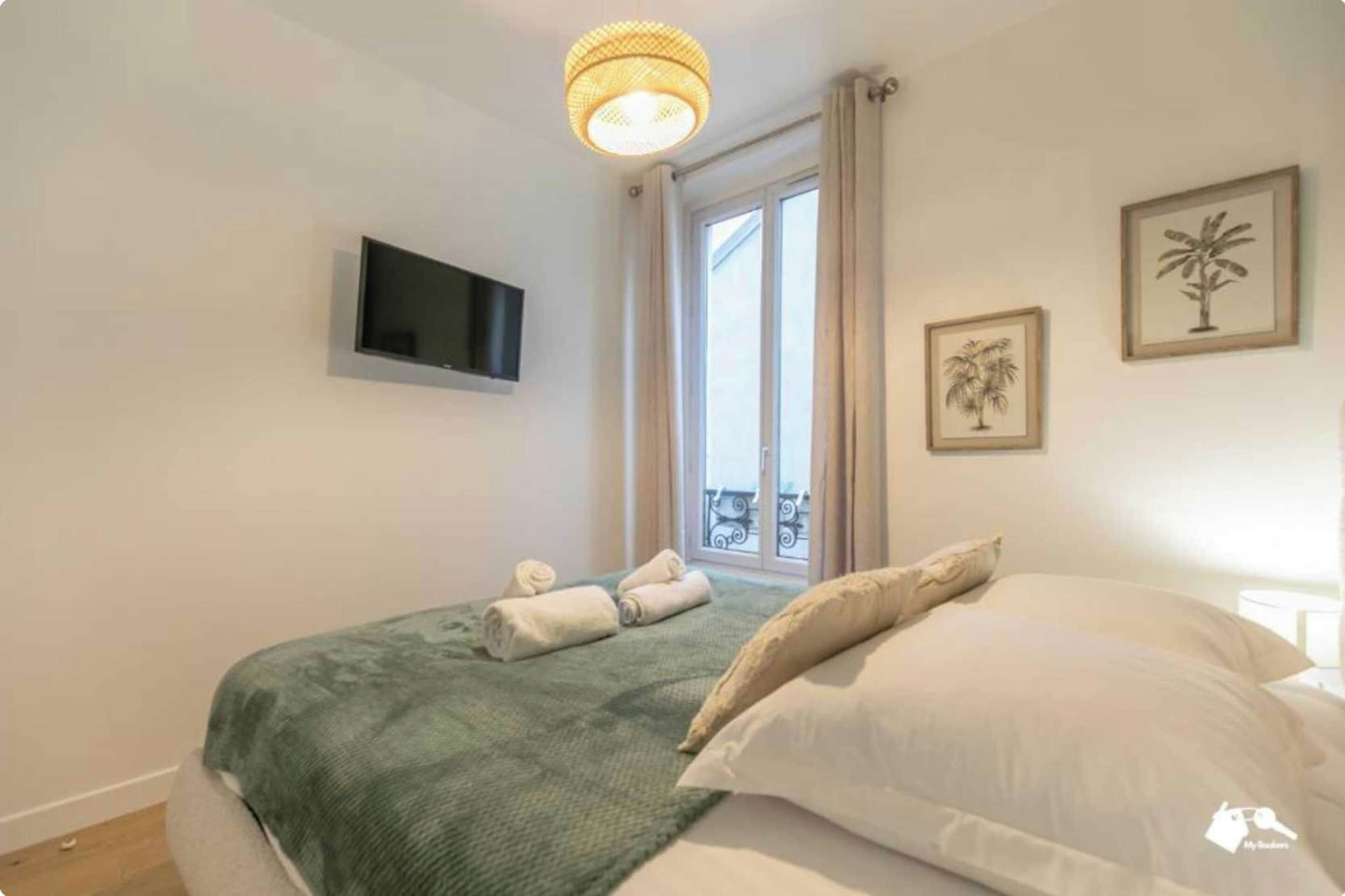 Une chambre à coucher avec un lit, un téléviseur fixé au mur, une fenêtre avec des rideaux et deux tableaux encadrés accrochés au mur.