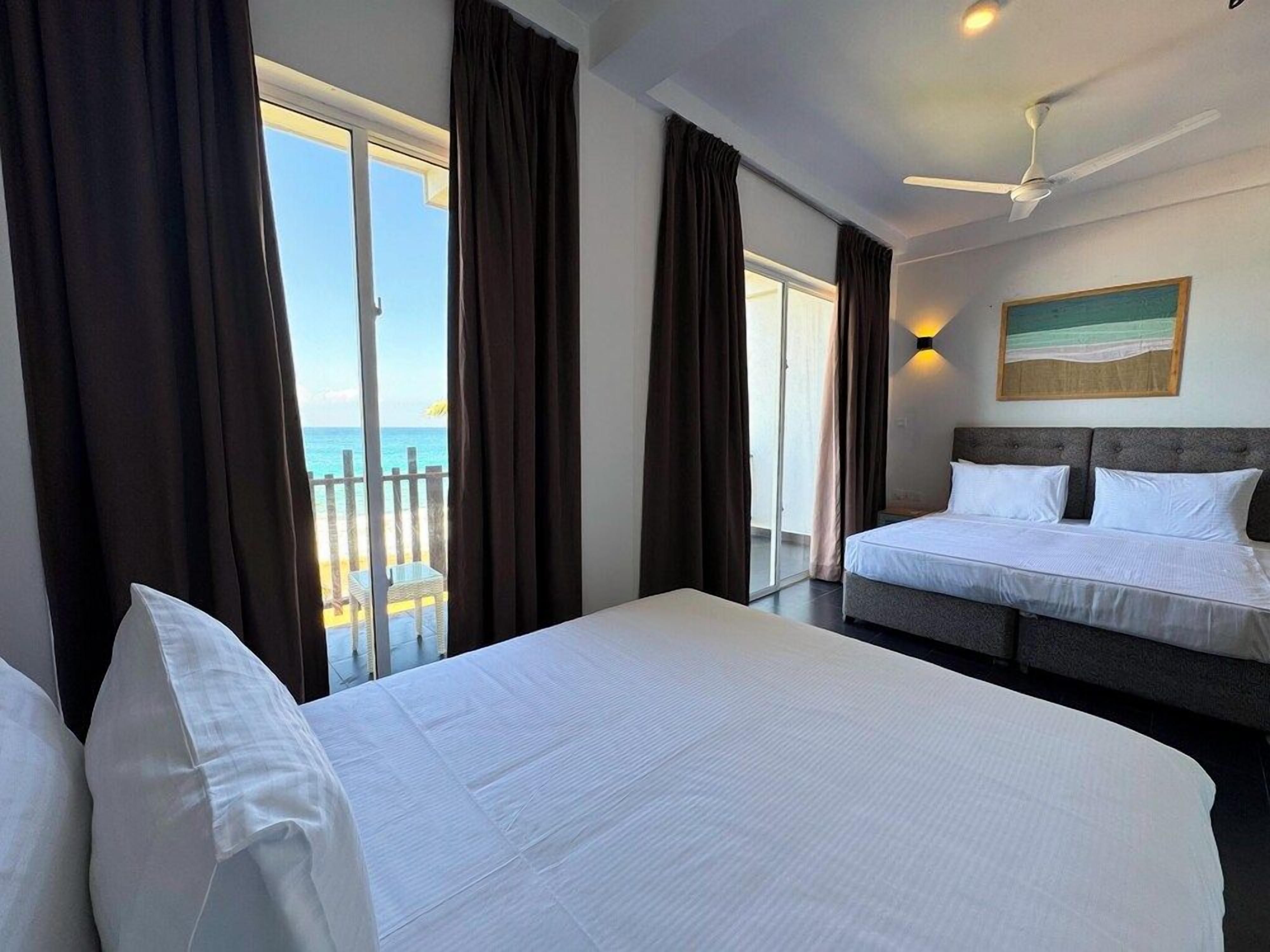 A hotel room with a bed, a ceiling fan, and a view of the sea through large windows.