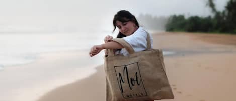 A person holding a tote bag with 'Moor Hotels' text, standing on a beach with sand and water.
