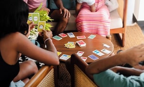 A group of people playing a card game around a round wooden table with cards and a small plant on it.