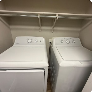 A laundry room with two front-loading washing machines and a metal shelf above. - Spacious 1bdr Townhome Minutes from ATT Stadium (Arlington)