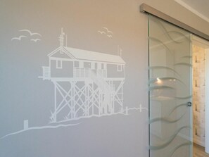 A wall with a white architectural drawing of a house on stilts, a glass door with a wavy pattern, and a wooden floor. - Utholm 5a - Kleine Brise (St. Peter-Ording)