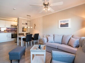 Una sala moderna con sofá, reposapiés y mesa de centro. Hay un comedor con sillas y una mesa. La habitación tiene un ventilador de techo y una cocina al fondo.