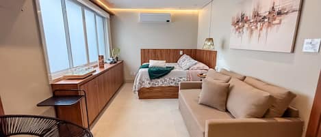 Habitación de hotel compacta con cama, sofá, mesita, silla y ventana con persianas.