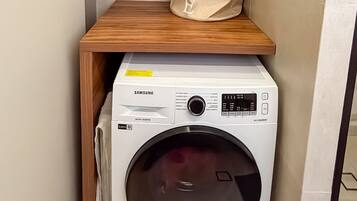 Un cuarto de lavandería con una lavadora Samsung digital, un gabinete de madera en la parte superior y una bolsa color beige con la etiqueta 'HOGAR' en un estante.