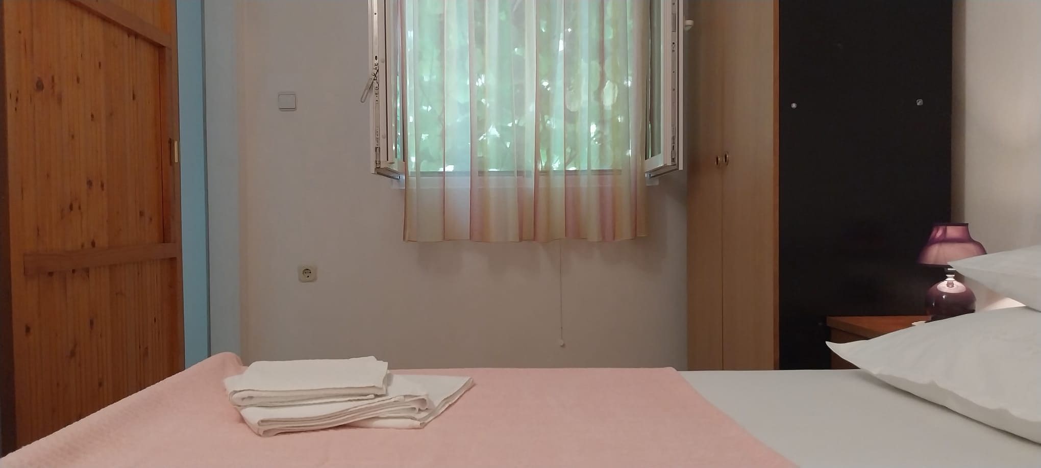 A room with a bed, a window with curtains, a door, and a nightstand with a lamp.