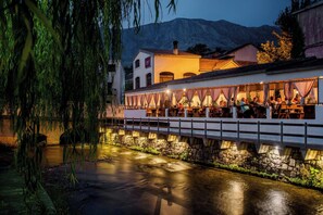 A riverside restaurant with outdoor seating overlooking a calm river, stone walls, and a mountain backdrop. - Kuča na rjeci s velikim vrtom I pogledom na planinu, 3 km.od mora-Split 10km. (Žrnovnica)