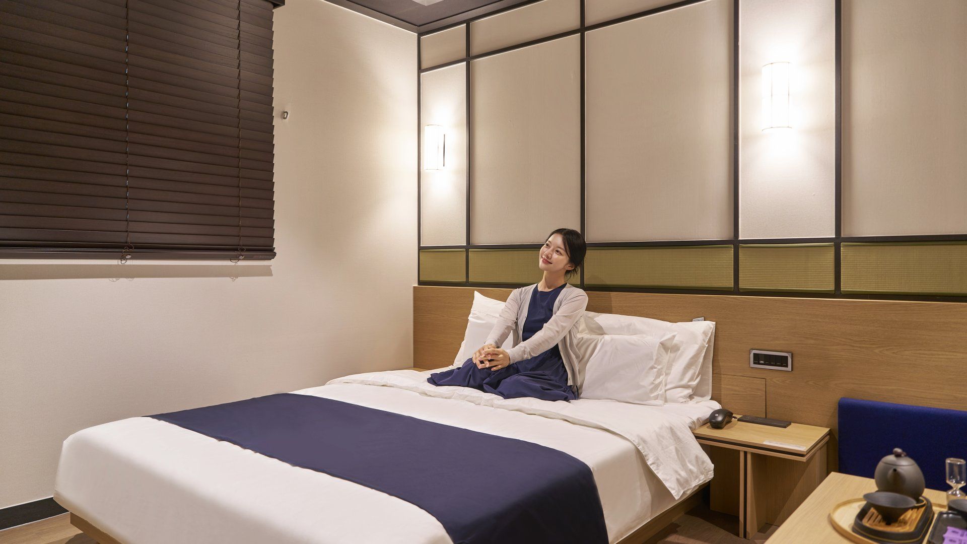 A hotel room with a bed, bedside table, and a window with blinds.