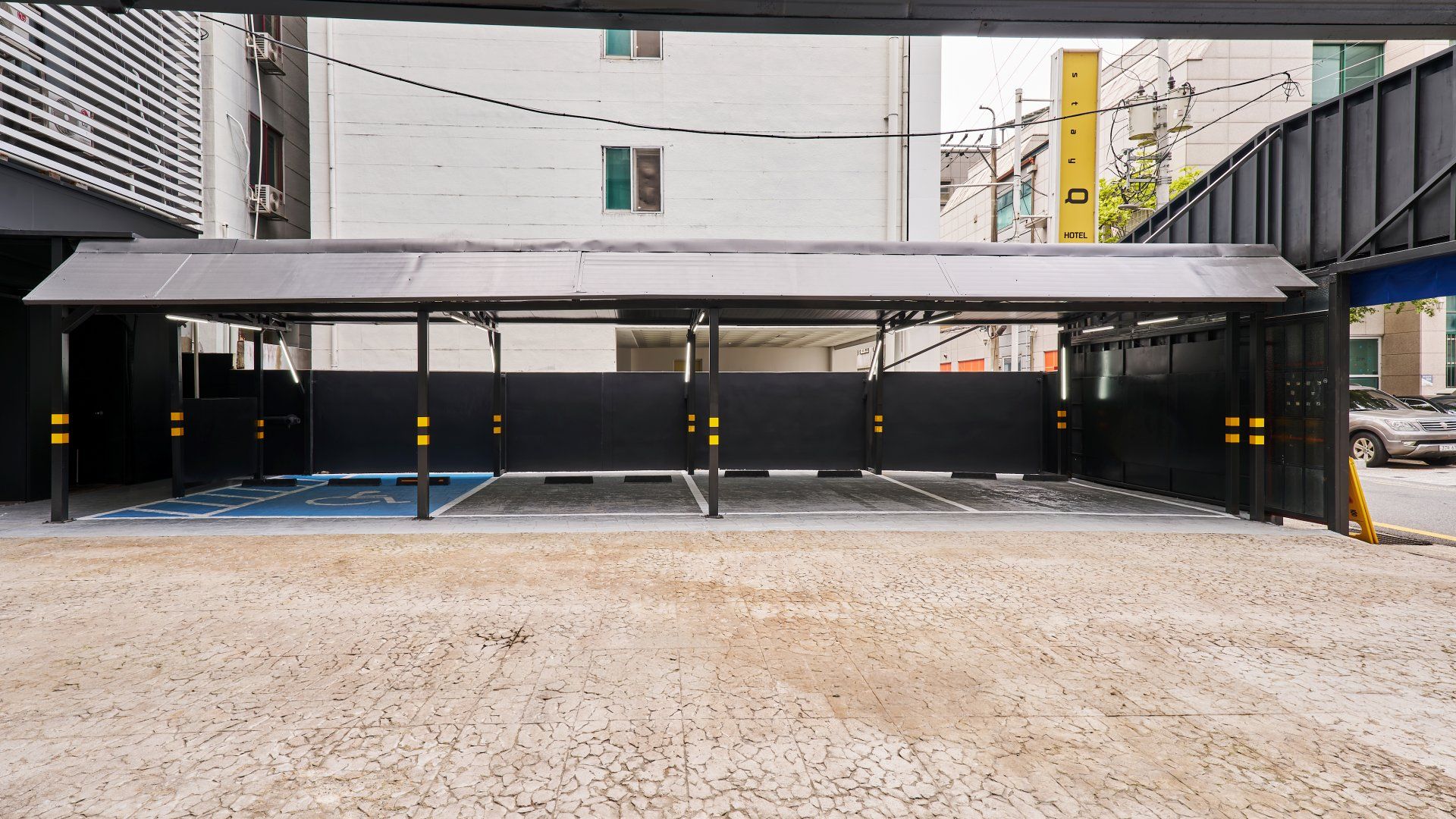 Un área de estacionamiento techada con un espacio reservado para discapacitados identificado con azul, un letrero de hotel amarillo y un edificio con fachada blanca al fondo.