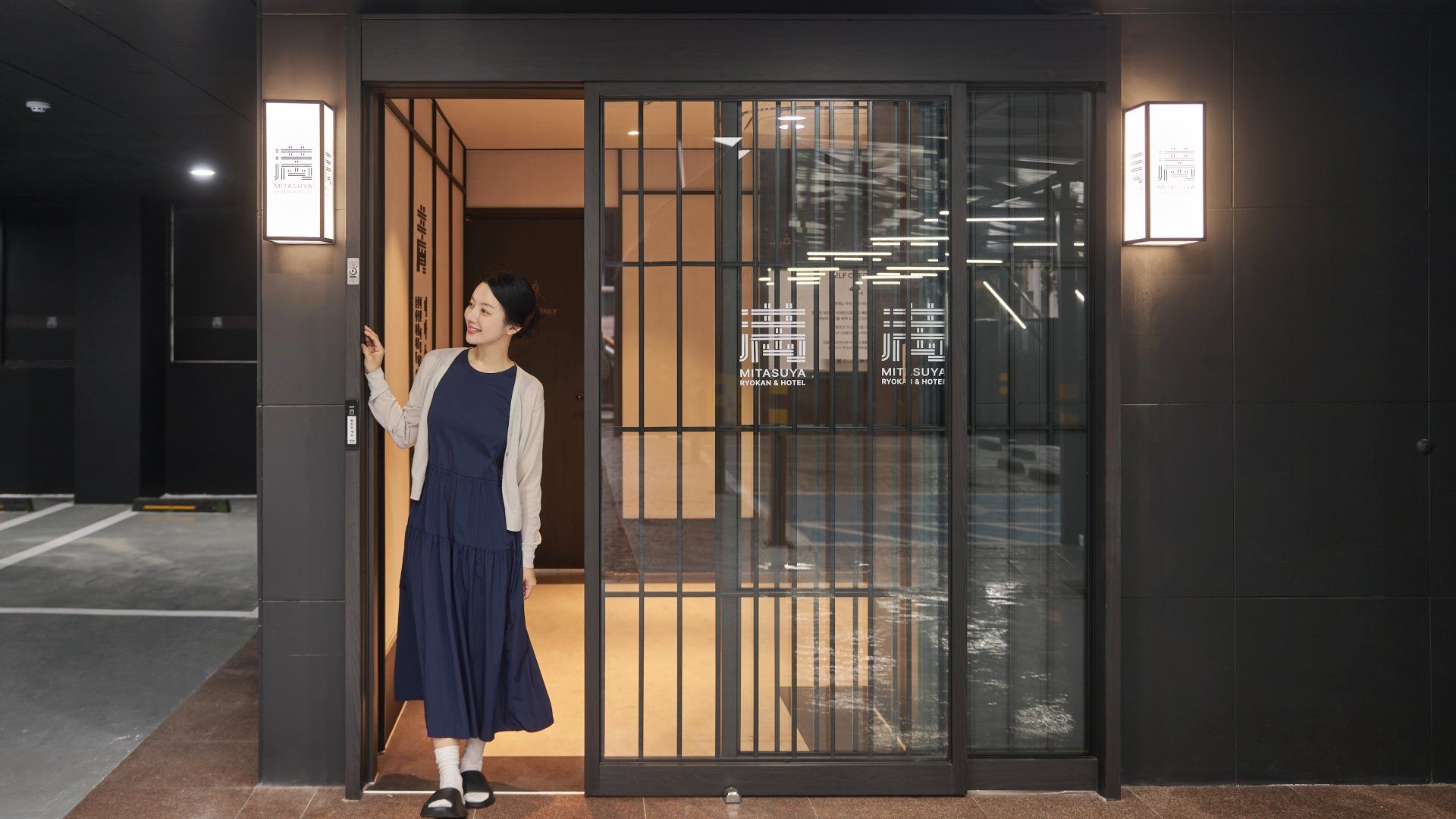 A person standing at the entrance of a building with a glass door, a sign with hotel branding, and a visible interior with modern lighting.
