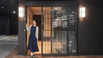 A person standing at the entrance of a building with a glass door, a sign with hotel branding, and a visible interior with modern lighting.