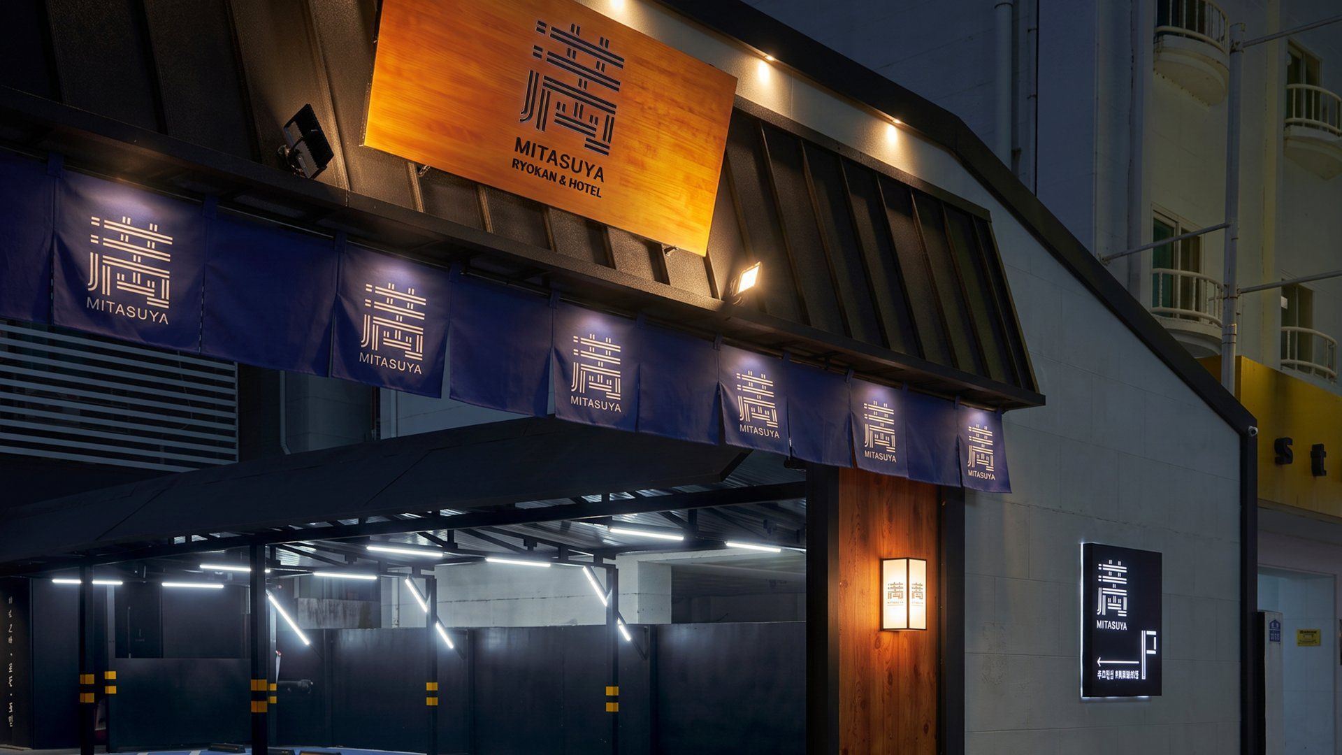 Entrada de un hotel con un letrero llamativo que dice 'MITSUYA RYOKAN & HOTEL' y varios banderines colgantes con el logotipo del hotel.