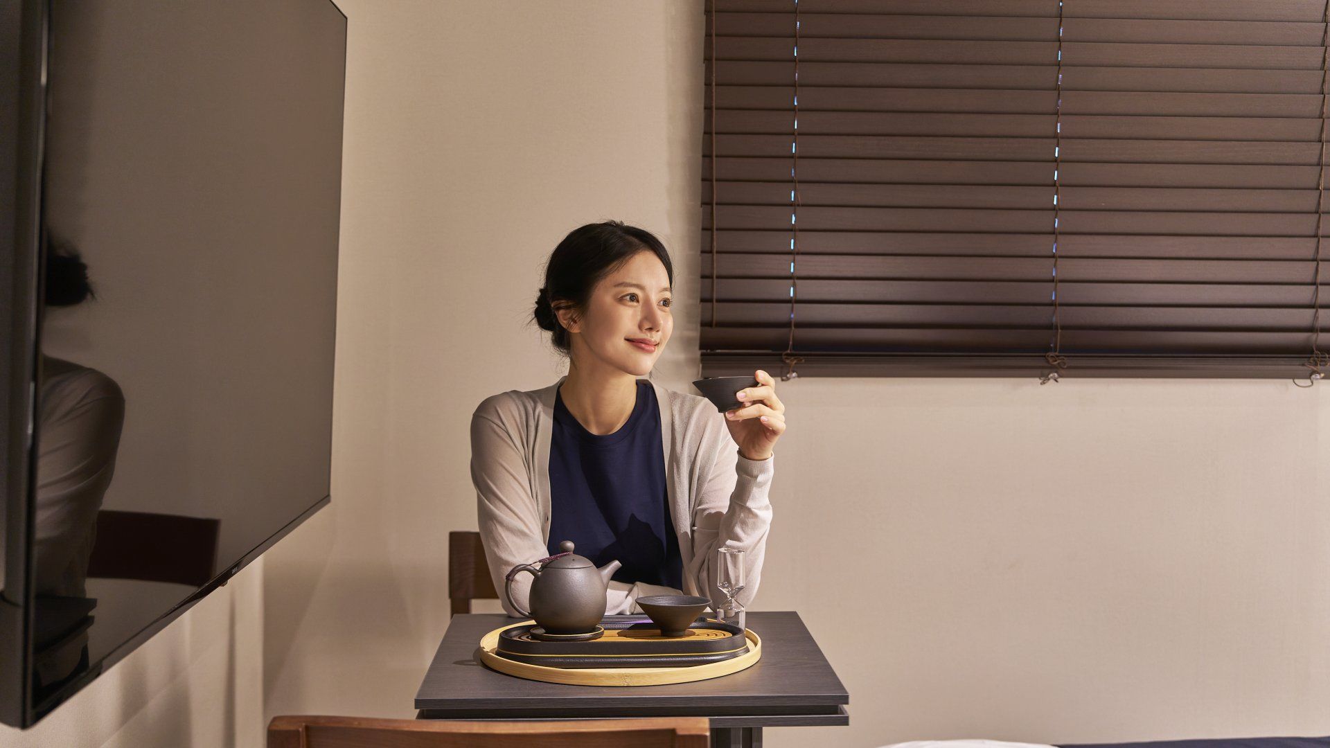 A person seated at a table with a teapot, cup, and saucer, with a window with blinds in the background.