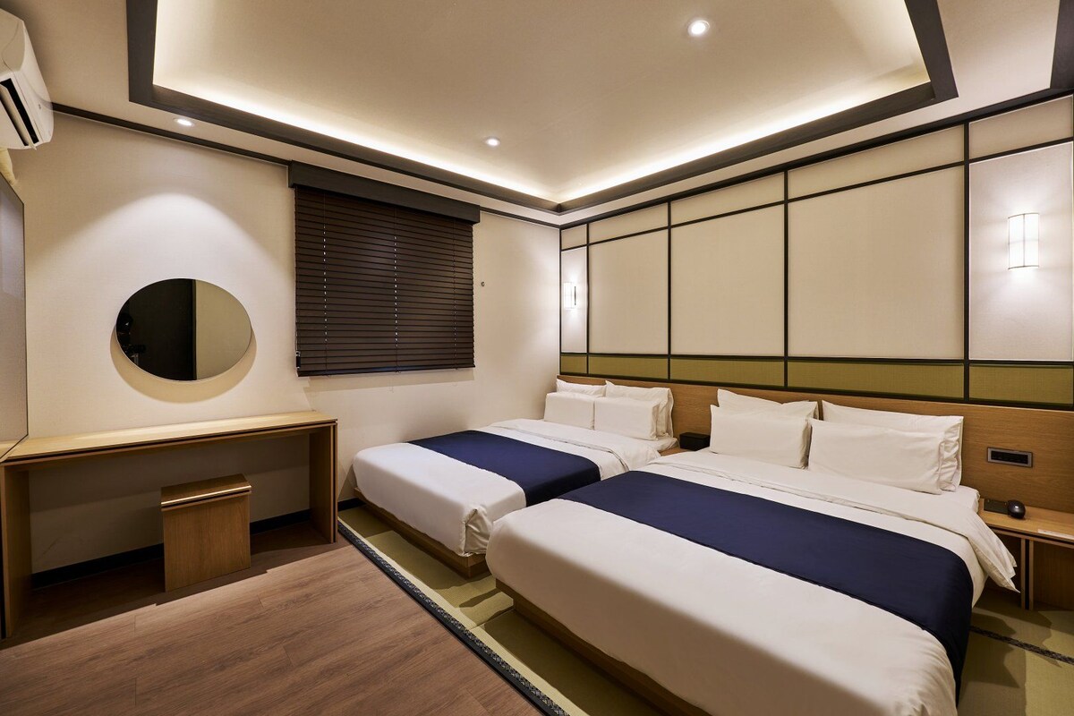 Habitación de hotel con dos camas, un escritorio, un espejo y una ventana con persianas.