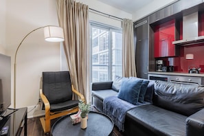 Ein modernes Wohnzimmer mit Sofa, Sessel, Stehlampe und Blick auf die Stadt durch große Fenster.