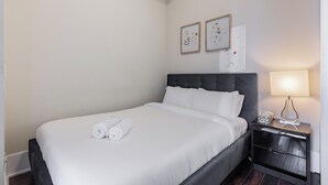 Una cama bien hecha con ropa blanca y toallas enrolladas, un cabecero oscuro, una mesita de noche con una lámpara y dos cuadros con motivos florales en la pared.