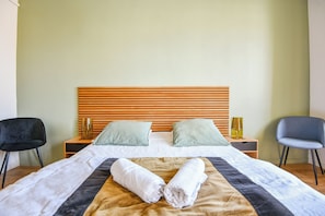 Quarto de hotel com cama, duas almofadas, mesa de cabeceira com abajur e duas cadeiras.