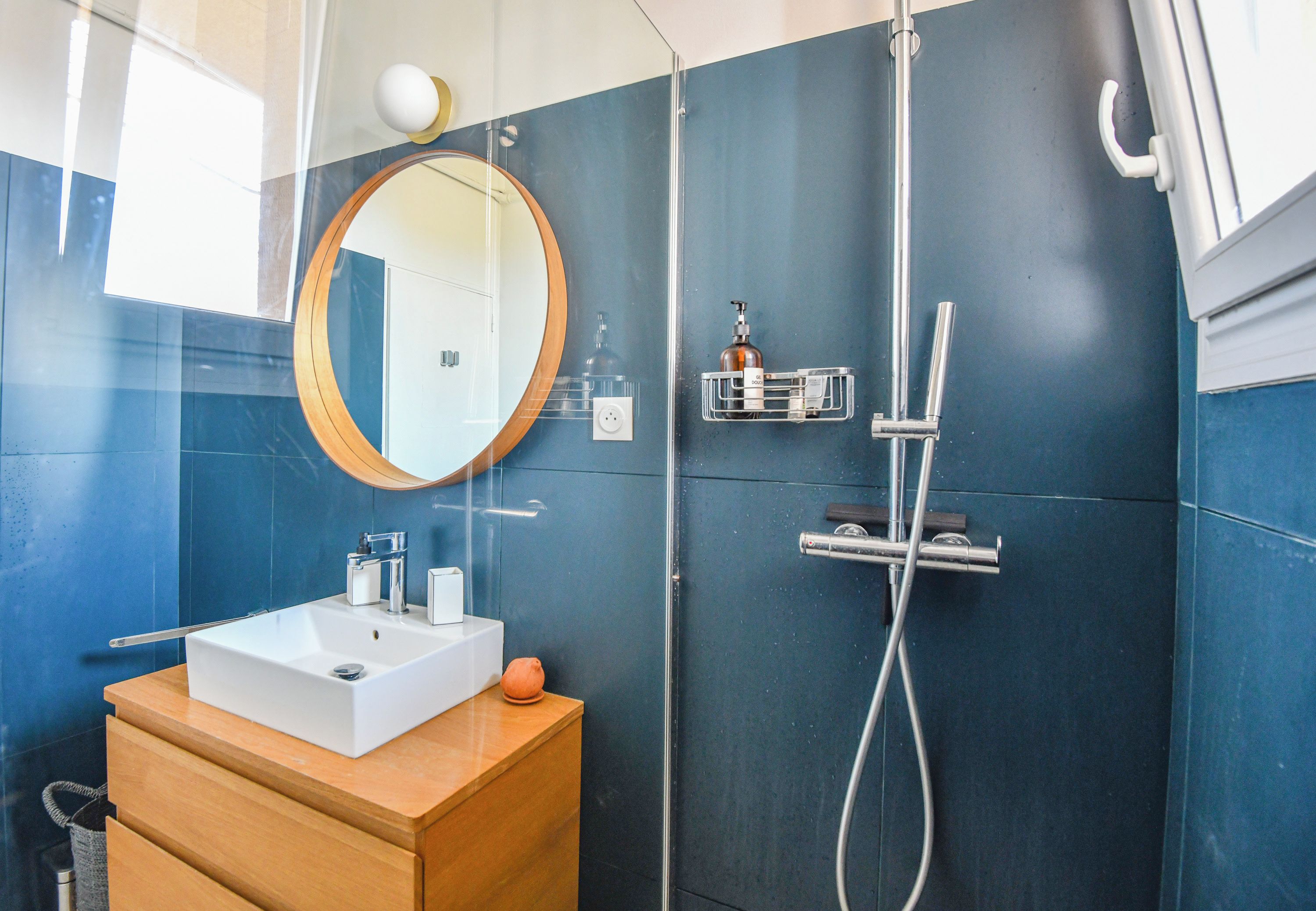 Un baño moderno con un lavabo blanco, tocador de madera, un espejo redondo y zona de ducha con cabezal de ducha manual.