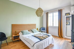 A hotel room with a bed, a chair, a bedside table, a lamp, a window with curtains, and a wall-mounted air conditioner. - Bail mobilité - Vue sur le Panier - Balcon - 2 ch (Marseille)