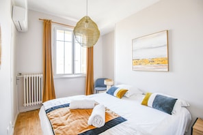 Ein Hotelzimmer mit Bett, einem Fenster mit Vorhängen, einem Heizkörper, einer Pendelleuchte, einem Sessel und einem Bild an der Wand.